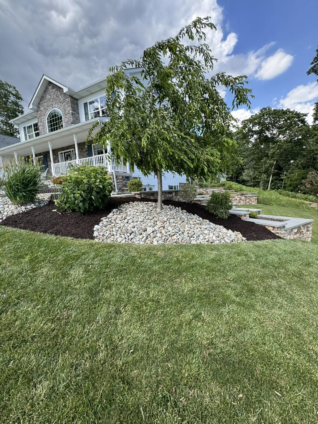 Freshly mulched landscape bed with river rock and a newly planted tree in front of a stone-and-siding home
